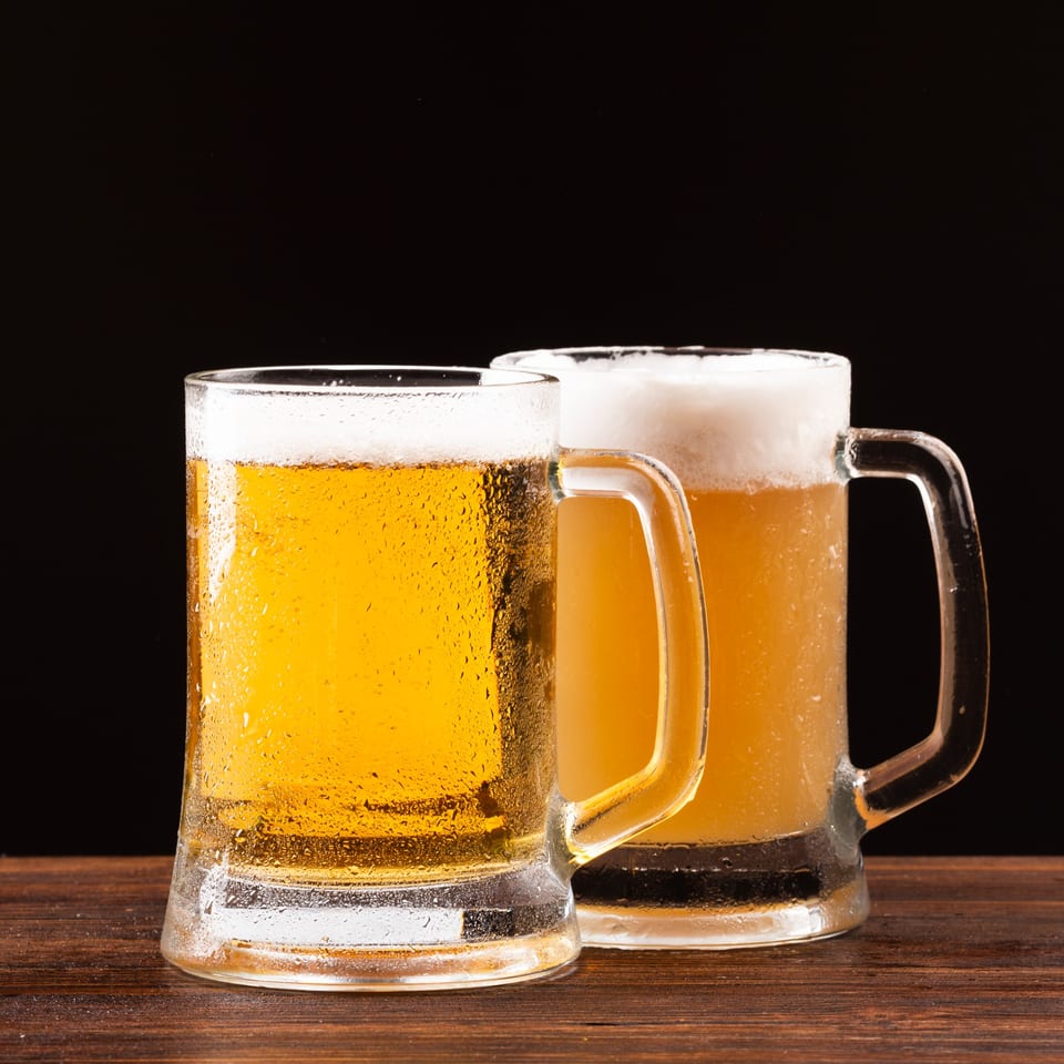 Two frosty mugs of golden beer with foamy heads sit on a dark wooden table.