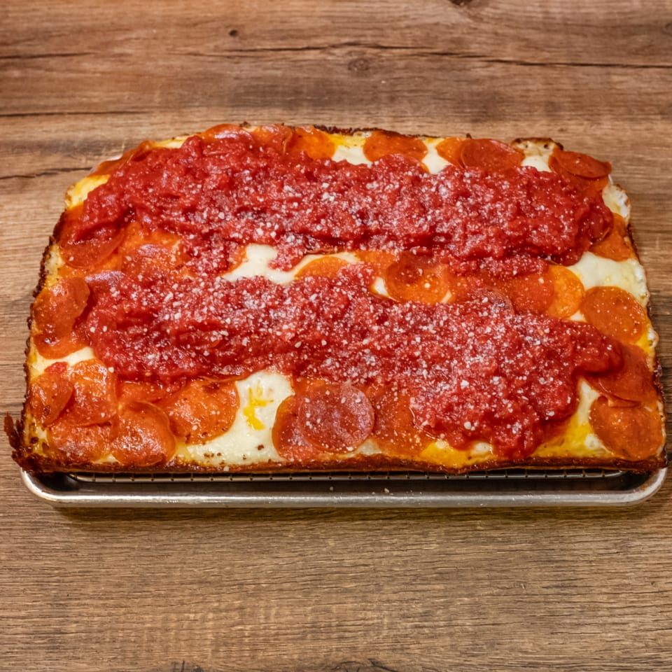 A rectangular Big Oven Pizza with pepperoni, cheese, and two stripes of red sauce on a metal rack.